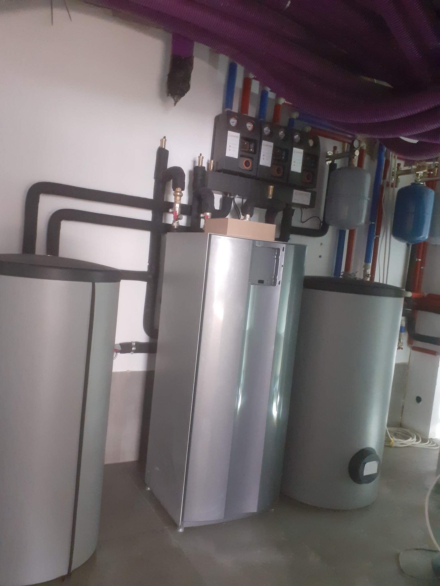 Instalacja zbiorników i kotła grzewczego w kotłowni w Bańskiej Niżnej z widocznymi rurami w kolorze fioletowym pod sufitem.