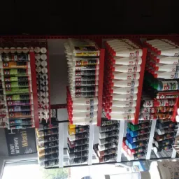 Półki regału w sklepie budowlanym wypełnione różnorodnymi materiałami wykończeniowymi i chemią budowlaną, w tym tuby z klejami, silikonami, piany montażowe w sprayu oraz pojemniki z płynami.