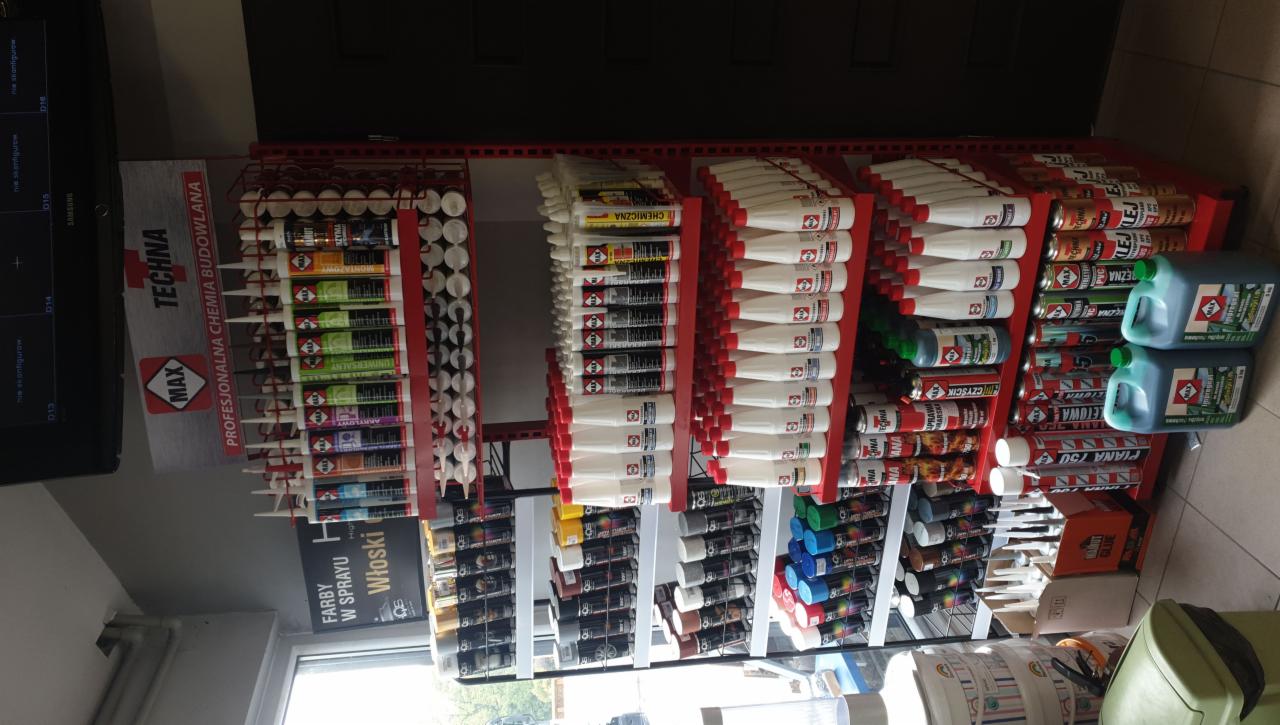 Półki regału w sklepie budowlanym wypełnione różnorodnymi materiałami wykończeniowymi i chemią budowlaną, w tym tuby z klejami, silikonami, piany montażowe w sprayu oraz pojemniki z płynami.
