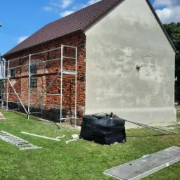 Budynek z częściowo otynkowaną elewacją, widoczne rusztowanie i materiały budowlane na trawie, ukazujące etap prac wykończeniowych.