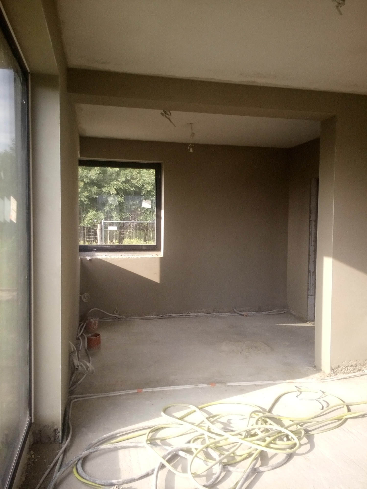 Wnętrze w stanie deweloperskim po tynkowaniu maszynowym, z widocznymi przewodami na podłodze i oknem z widokiem na zieleń.