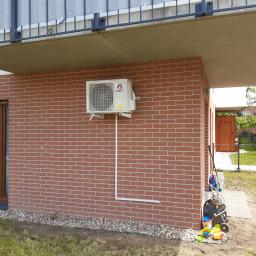 Mikart - Zewnętrzna jednostka klimatyzatora marki Gree zamontowana na ceglanej ścianie domu jednorodzinnego, z widocznymi przewodami i elementami instalacji, obok okna i zabawek ogrodowych.