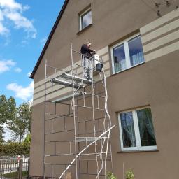 Mikart - Mężczyzna w trakcie montażu jednostki zewnętrznej klimatyzacji na rusztowaniu przy brązowym domu z białymi oknami, widoczne kable i pudełko z nowym urządzeniem na dole.