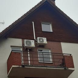 Mikart - Dwa klimatyzatory zamontowane na elewacji budynku z balkonem, widoczne rury odprowadzające skropliny, pochmurne niebo w tle.