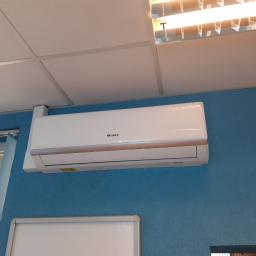 Mikart - Biały klimatyzator marki Gree zamontowany na niebieskiej, teksturowanej ścianie w biurze, widok z dołu, w tle biały sufit kasetonowy i świetlówka.