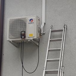 Mikart - Zewnętrzna jednostka klimatyzacyjna GREE zamontowana na szarej, chropowatej ścianie budynku, z podłączonym manometrem i widoczną drabiną opartą o ścianę.