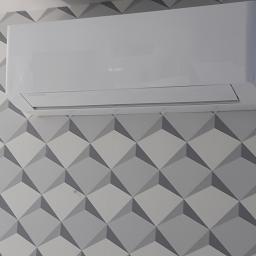 Mikart - Biała jednostka klimatyzacyjna marki Gree zamontowana na ścianie z geometryczną tapetą w odcieniach szarości.