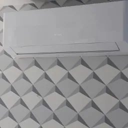 Biała jednostka klimatyzacyjna marki Gree zamontowana na ścianie z geometryczną tapetą w odcieniach szarości.