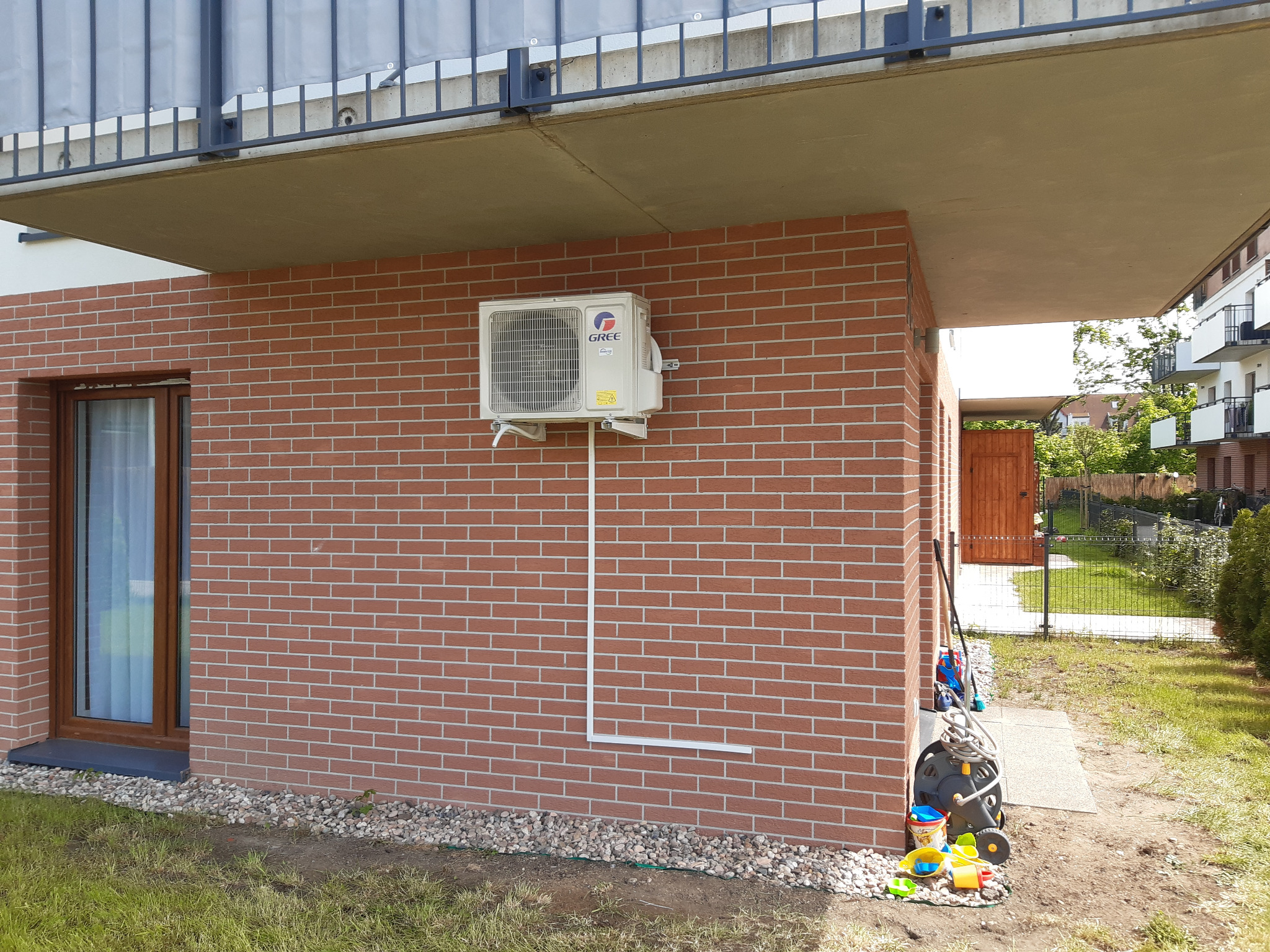 Zewnętrzna jednostka klimatyzatora marki Gree zamontowana na ceglanej ścianie domu jednorodzinnego, z widocznymi przewodami i elementami instalacji, obok okna i zabawek ogrodowych.