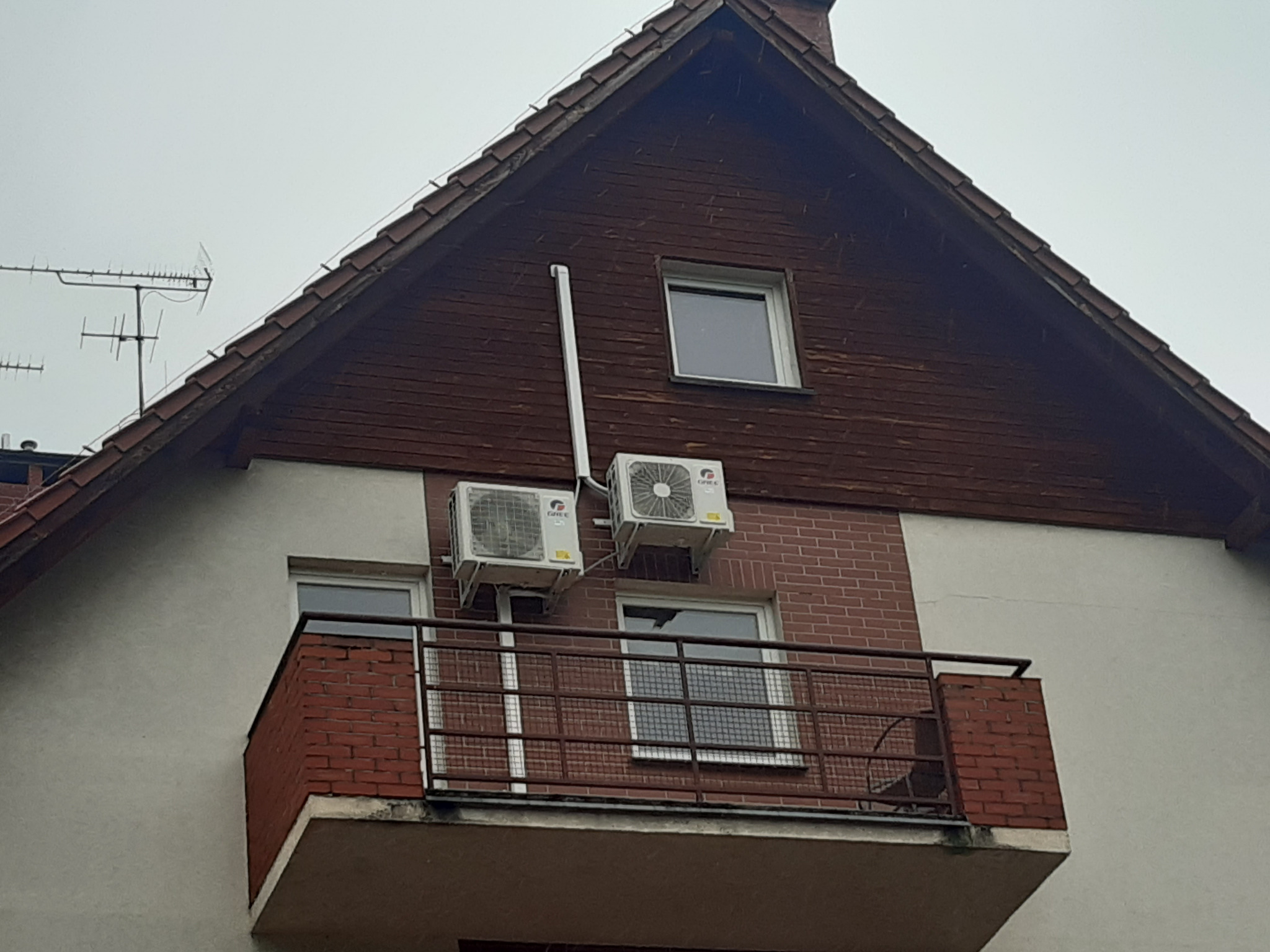 Dwa klimatyzatory zamontowane na elewacji budynku z balkonem, widoczne rury odprowadzające skropliny, pochmurne niebo w tle.