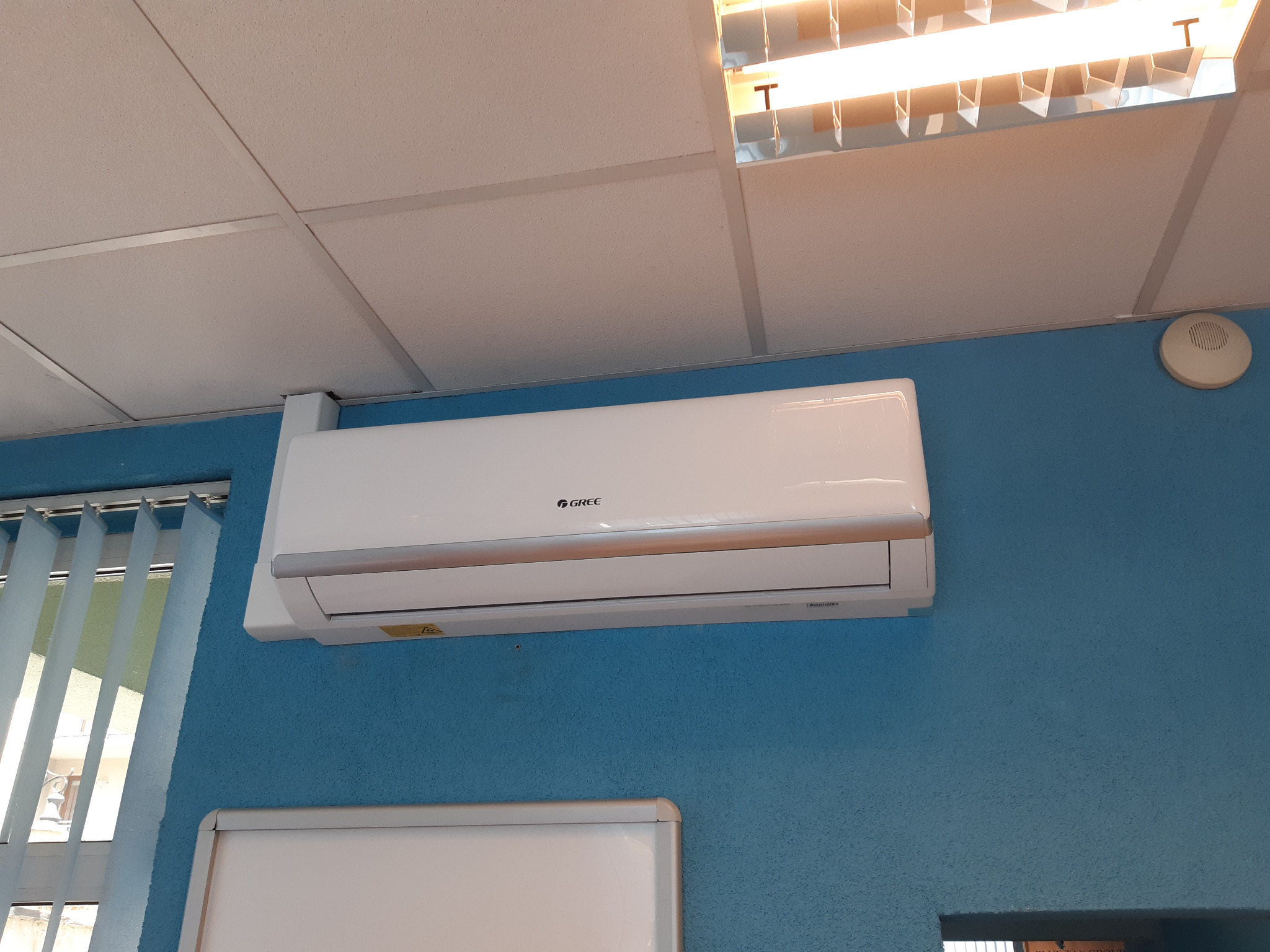 Biały klimatyzator marki Gree zamontowany na niebieskiej, teksturowanej ścianie w biurze, widok z dołu, w tle biały sufit kasetonowy i świetlówka.