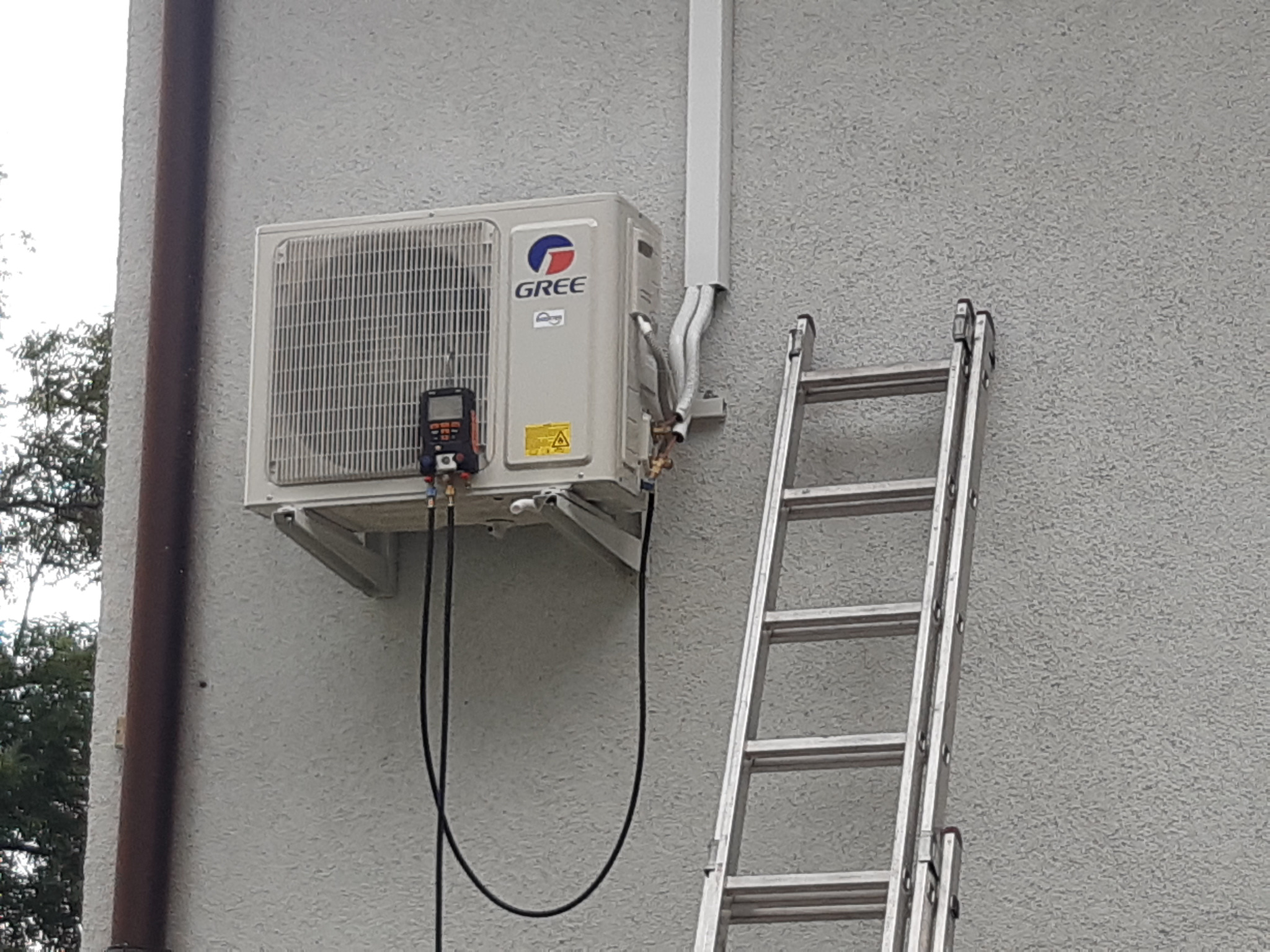 Zewnętrzna jednostka klimatyzacyjna GREE zamontowana na szarej, chropowatej ścianie budynku, z podłączonym manometrem i widoczną drabiną opartą o ścianę.