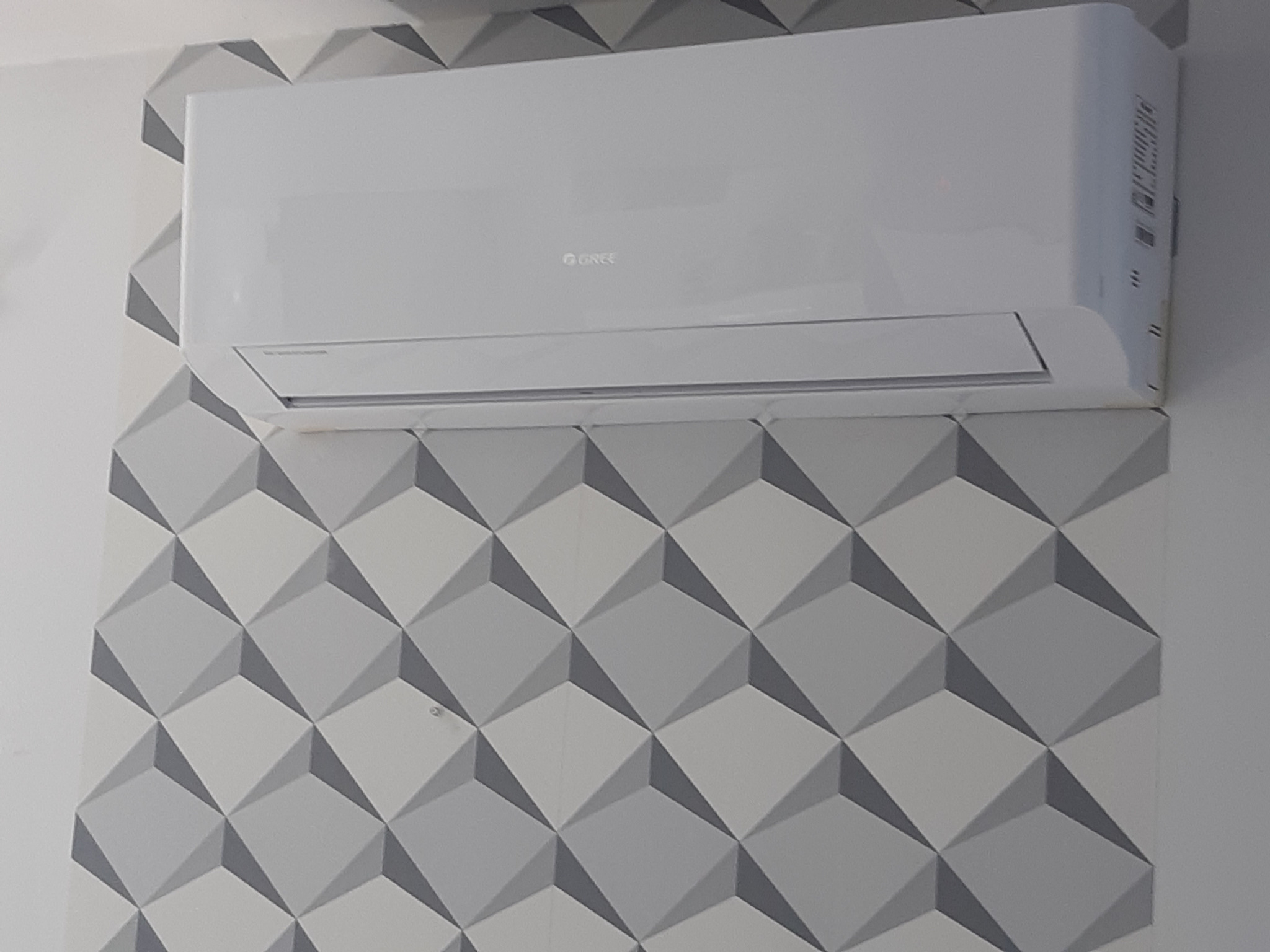 Biała jednostka klimatyzacyjna marki Gree zamontowana na ścianie z geometryczną tapetą w odcieniach szarości.