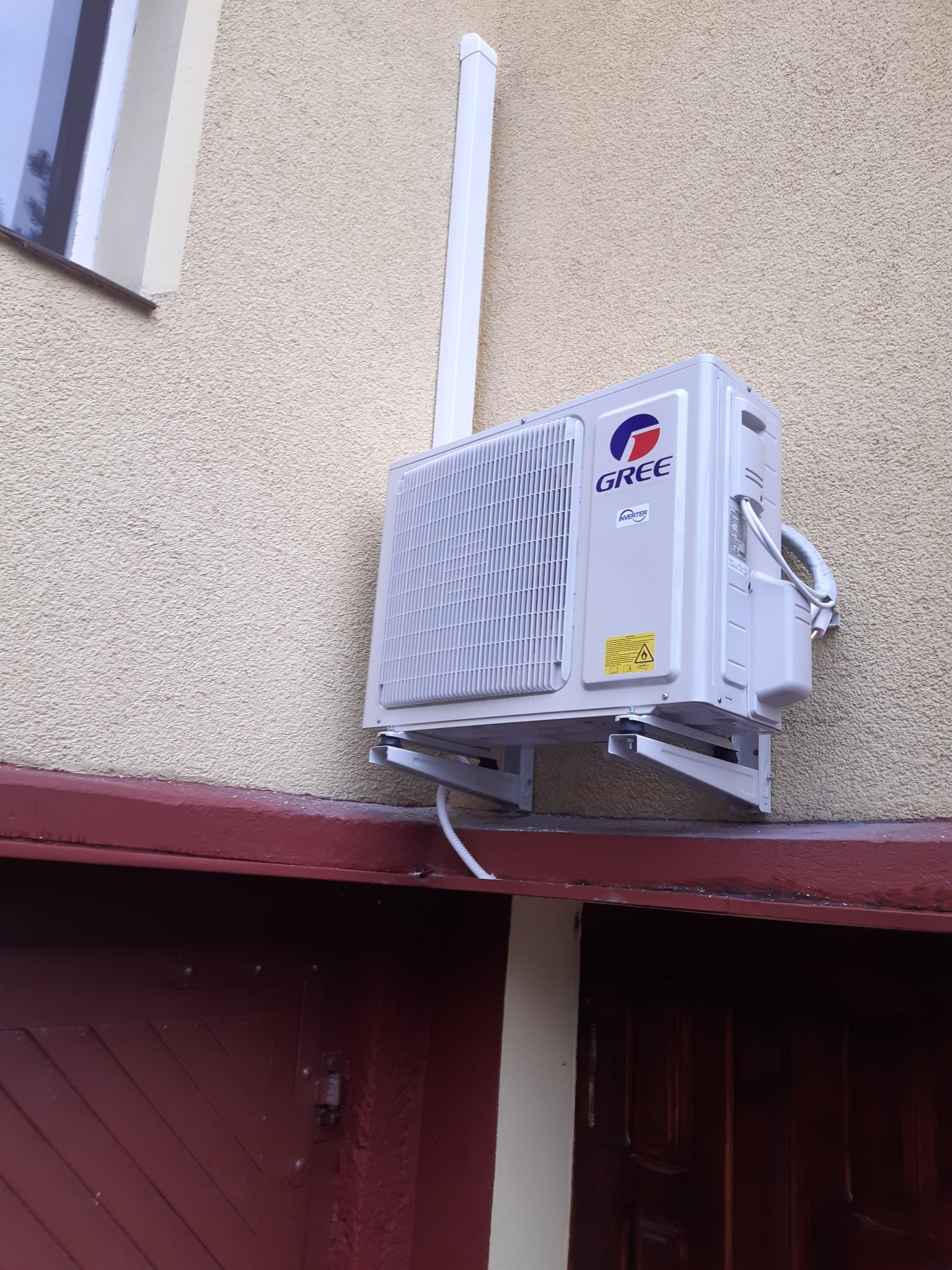 Zewnętrzna jednostka klimatyzatora marki Gree zamontowana na elewacji budynku, widoczna rura odprowadzająca skropliny i przewody.