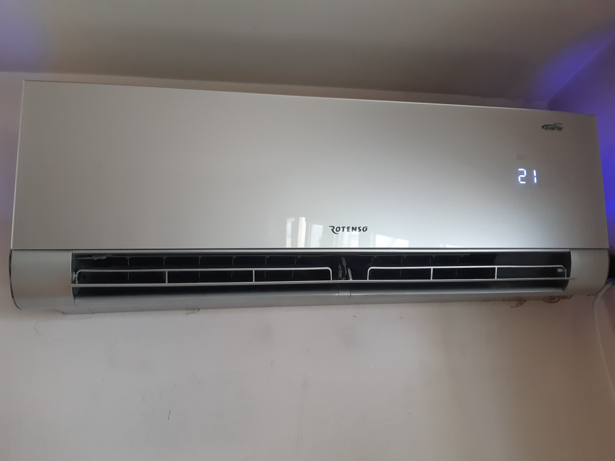 Zamontowany na ścianie, biały klimatyzator Rotenso z widocznym wyświetlaczem temperatury 21 stopni Celsjusza.