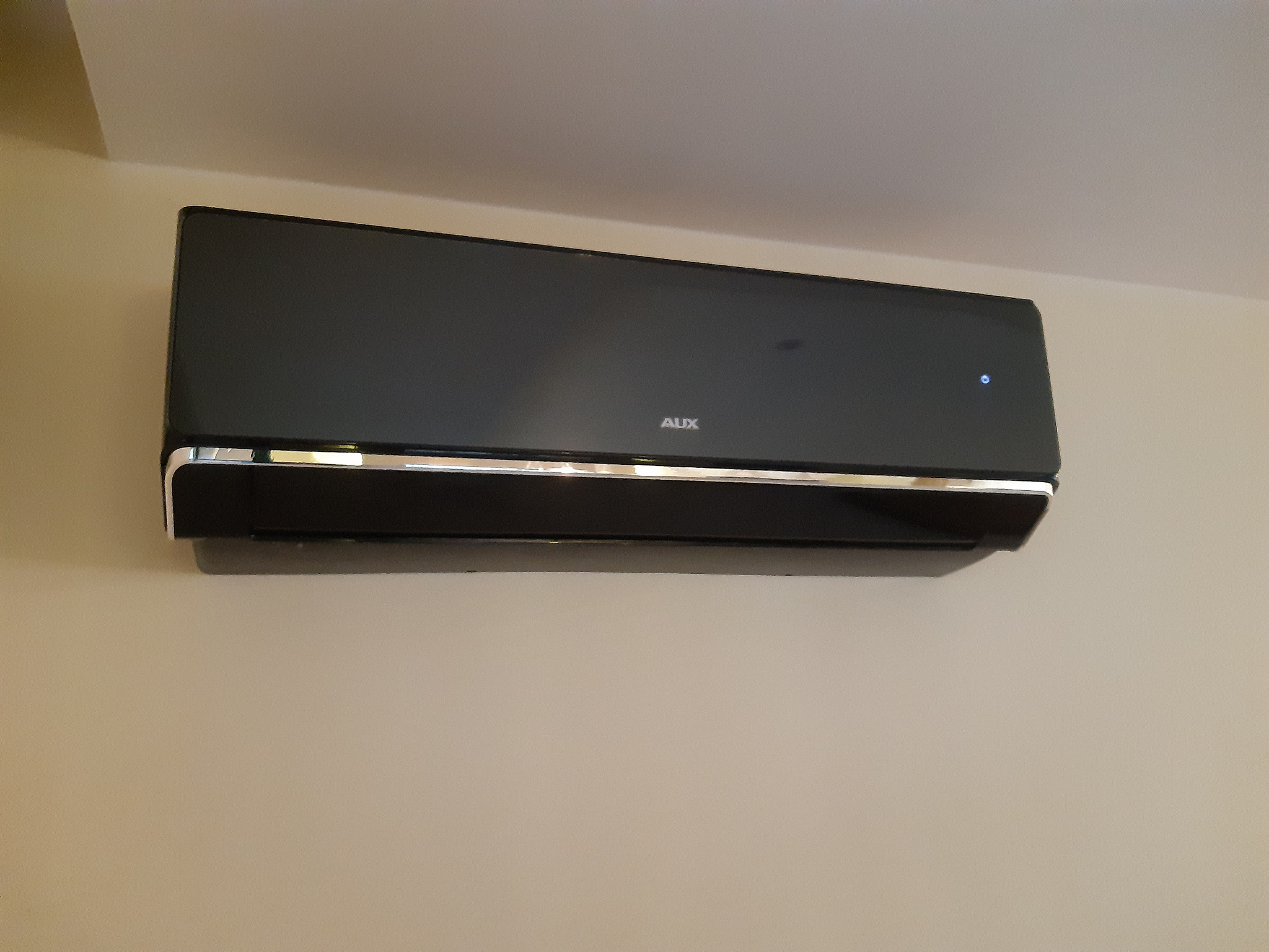 Czarny klimatyzator marki AUX zamontowany na beżowej ścianie, widoczna delikatna srebrna listwa i świecąca dioda.