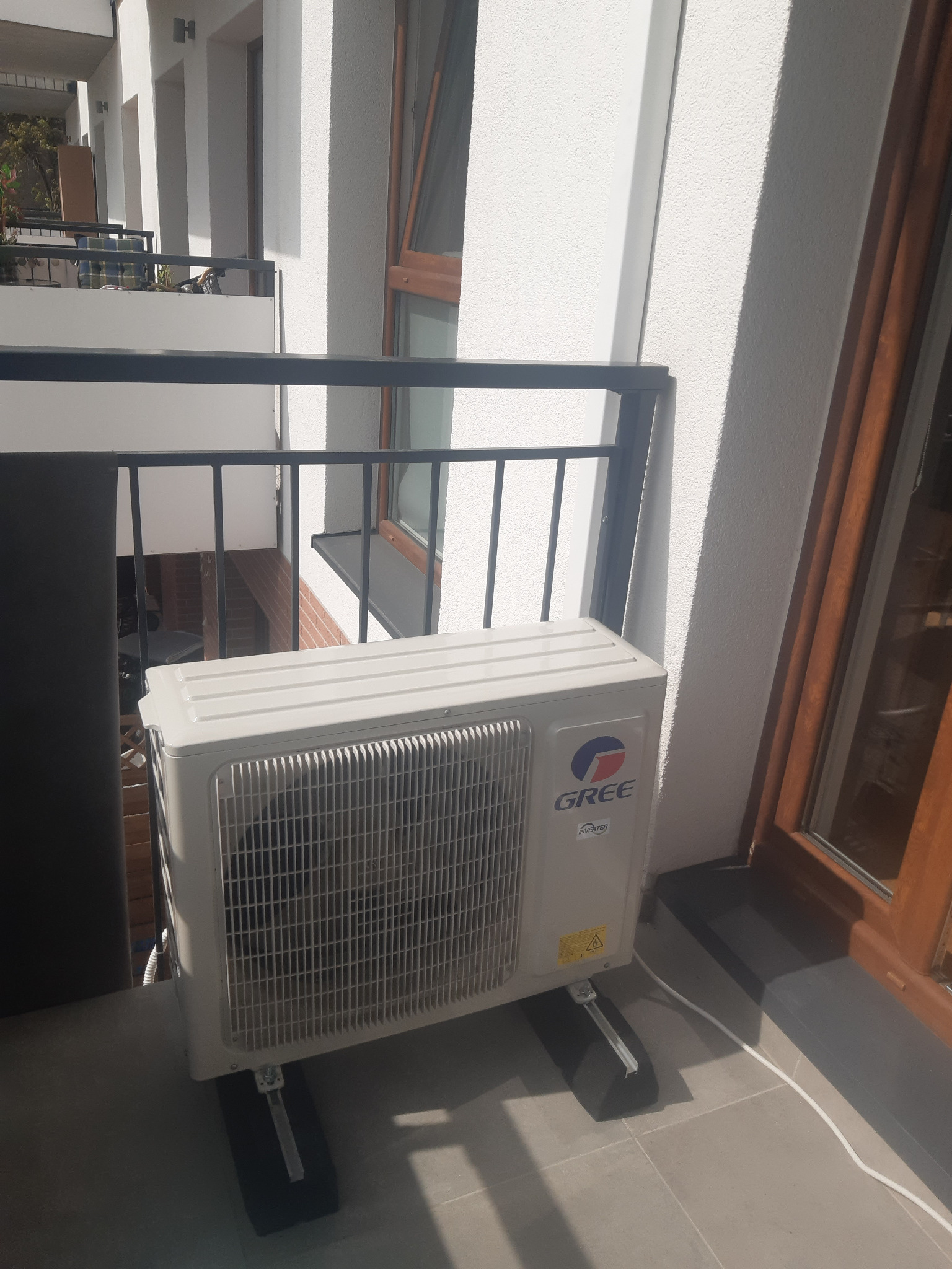 Zewnętrzna jednostka klimatyzacji marki Gree zamontowana na balkonie apartamentu, widoczne wsporniki antywibracyjne oraz kabel zasilający.