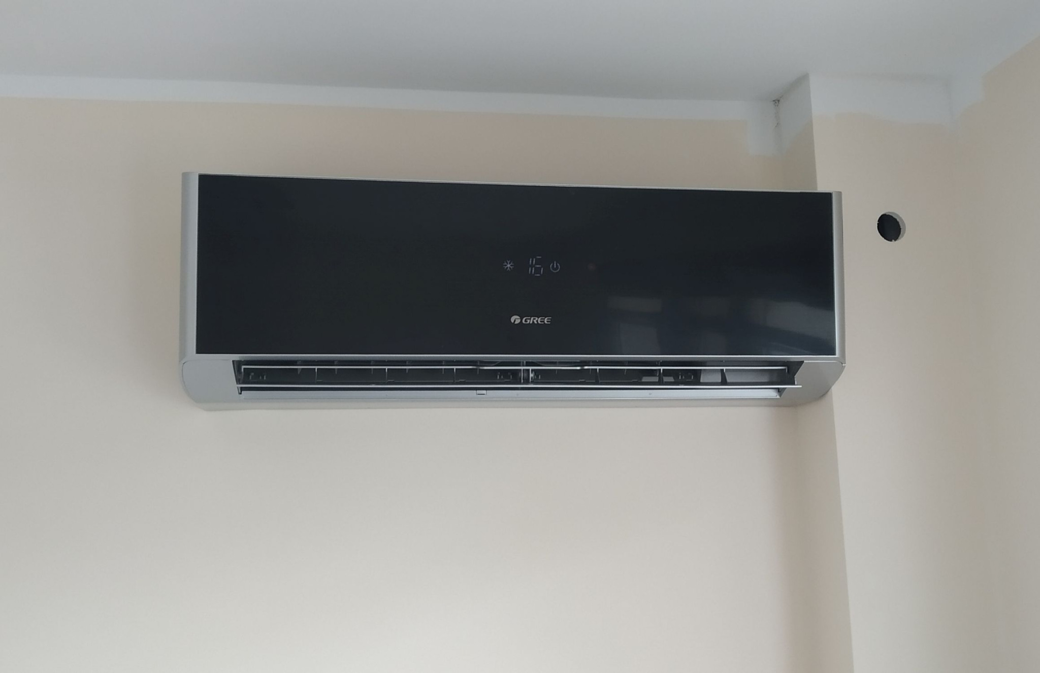 Czarny klimatyzator marki Gree zamontowany na beżowej ścianie, widoczne niedomalowane fragmenty sufitu i ściany w rogu pomieszczenia oraz otwór w ścianie po lewej stronie klimatyzatora.