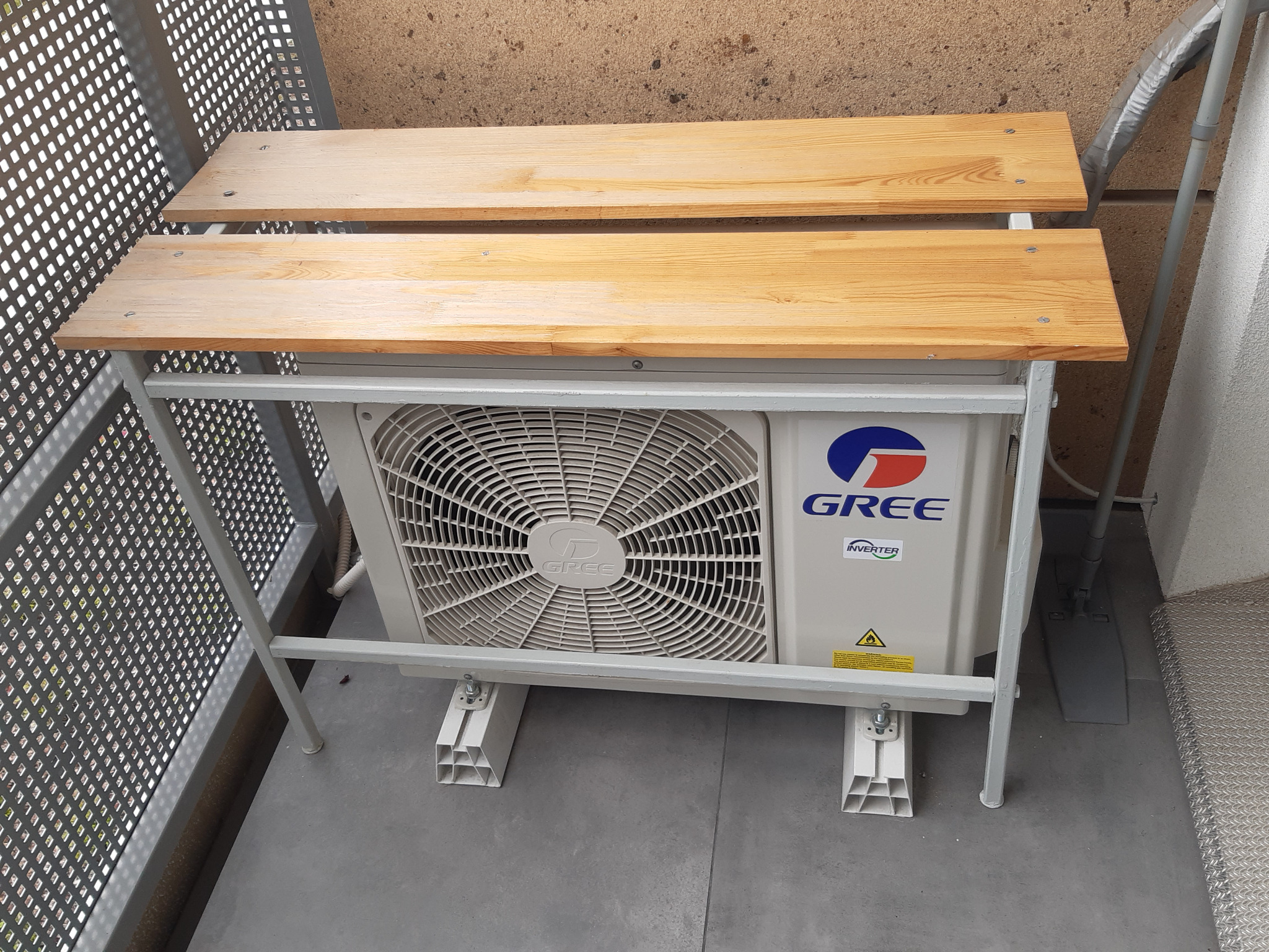 Zewnętrzna jednostka klimatyzacji GREE zamontowana na metalowym stelażu z drewnianymi blatami, stojąca na balkonie.
