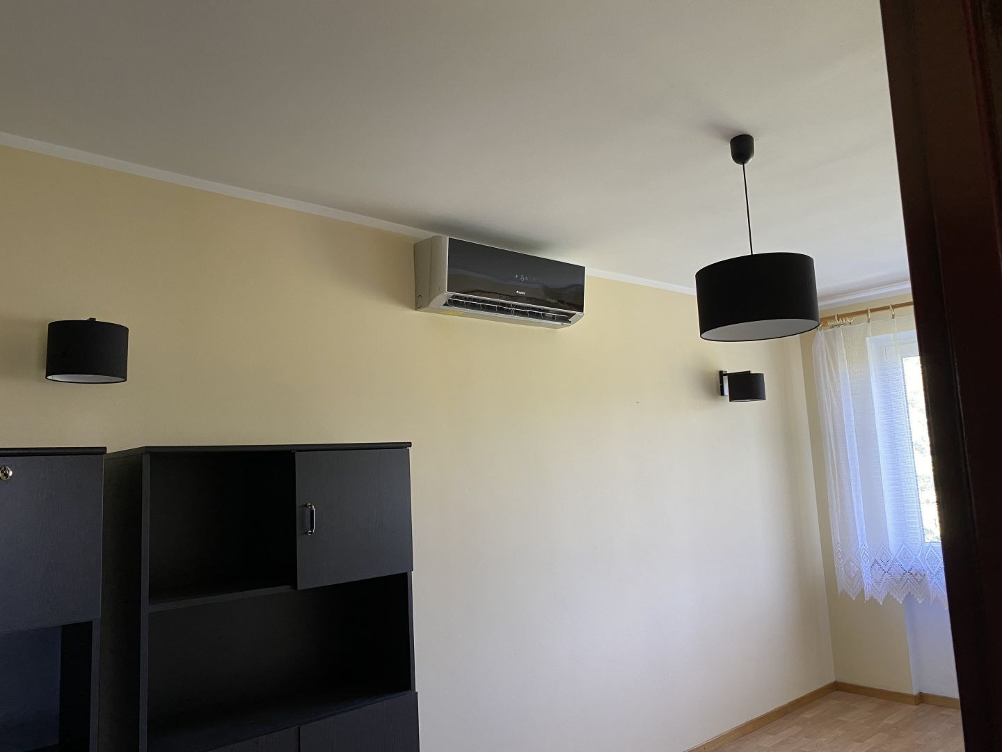Wnętrze pokoju z zamontowanym klimatyzatorem ściennym, czarnymi lampami i meblami w kolorze grafitowym, widok na okno z firanką.