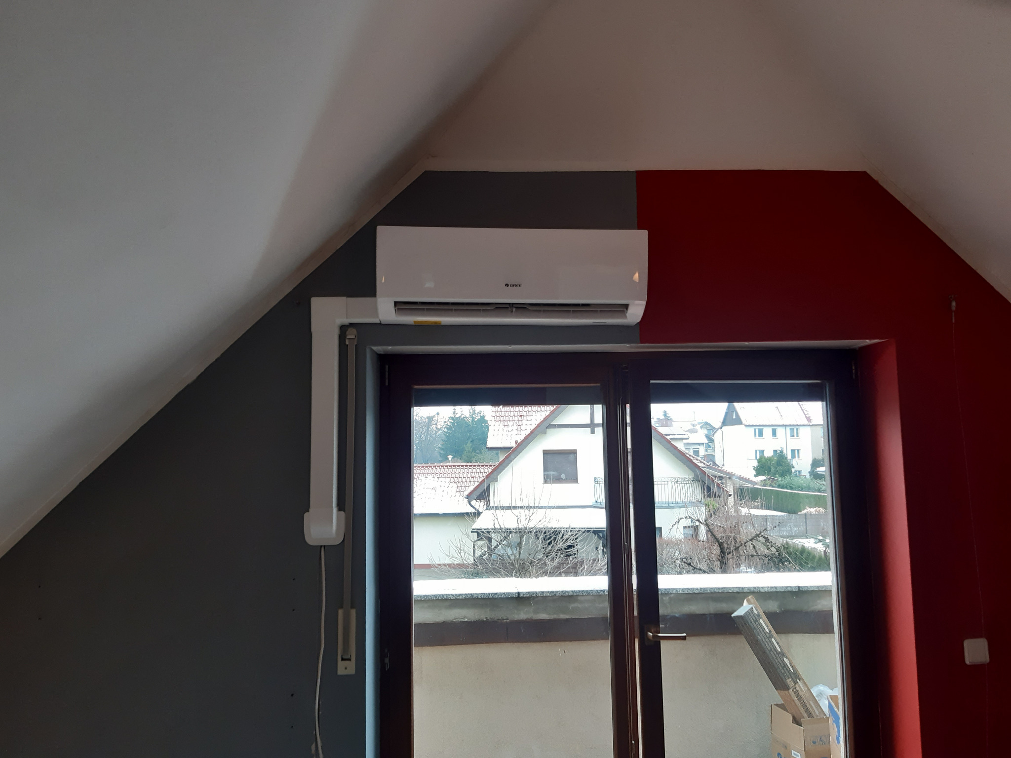 Biały klimatyzator marki Gree zamontowany na ścianie pod skosem w pokoju na poddaszu, widok z okna na ośnieżone dachy domów.