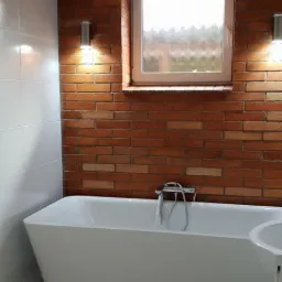 Nowoczesna wanna wolnostojąca z baterią, umieszczona na tle ściany z cegły i białych płytek w łazience w Pabianicach, oświetlona przez dwa kinkiety po bokach okna.