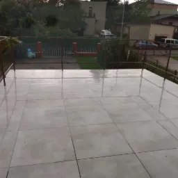 Wyłożony jasnymi płytkami taras z metalową balustradą w pochmurny dzień, widok na sąsiednie domy i zieleń.