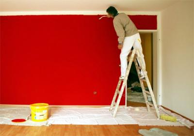 Mężczyzna maluje intensywnie czerwoną ścianę z drabiny, żółte wiadro farby stoi na podłodze zabezpieczonej folią. Widoczne białe obramowanie i fragment sąsiedniego pokoju.