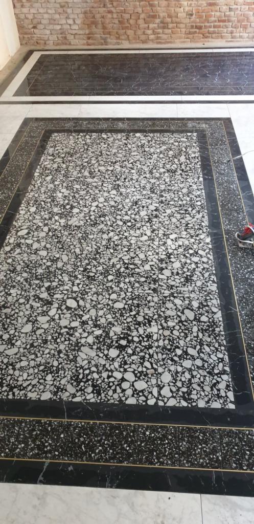 Wykończona podłoga z płytek lastryko w czarno-białej kolorystyce, otoczona czarnymi płytkami z delikatnym złotym akcentem, na tle fragmentu ściany z cegły.