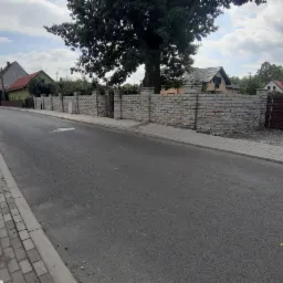 Długi, łukowaty mur z jasnego kamienia, oddzielający posesję od ulicy, z widocznym drzewem i zabudowaniami w tle.