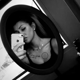 Czarno-białe selfie kobiety w owalnym lustrze, z telefonem iPhone w dłoni, widoczne tatuaże na dekolcie, okno w tle. Ujęcie z góry, lekko obrócone.