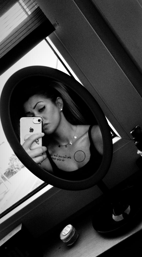 Czarno-białe selfie kobiety w owalnym lustrze, z telefonem iPhone w dłoni, widoczne tatuaże na dekolcie, okno w tle. Ujęcie z góry, lekko obrócone.