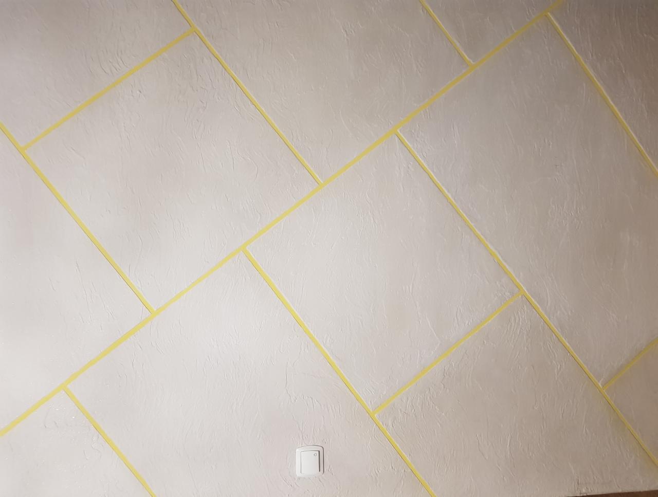 Ściana z nałożoną strukturą i wyklejonymi taśmami malarskimi tworzącymi geometryczny wzór przed malowaniem, widoczny włącznik światła.