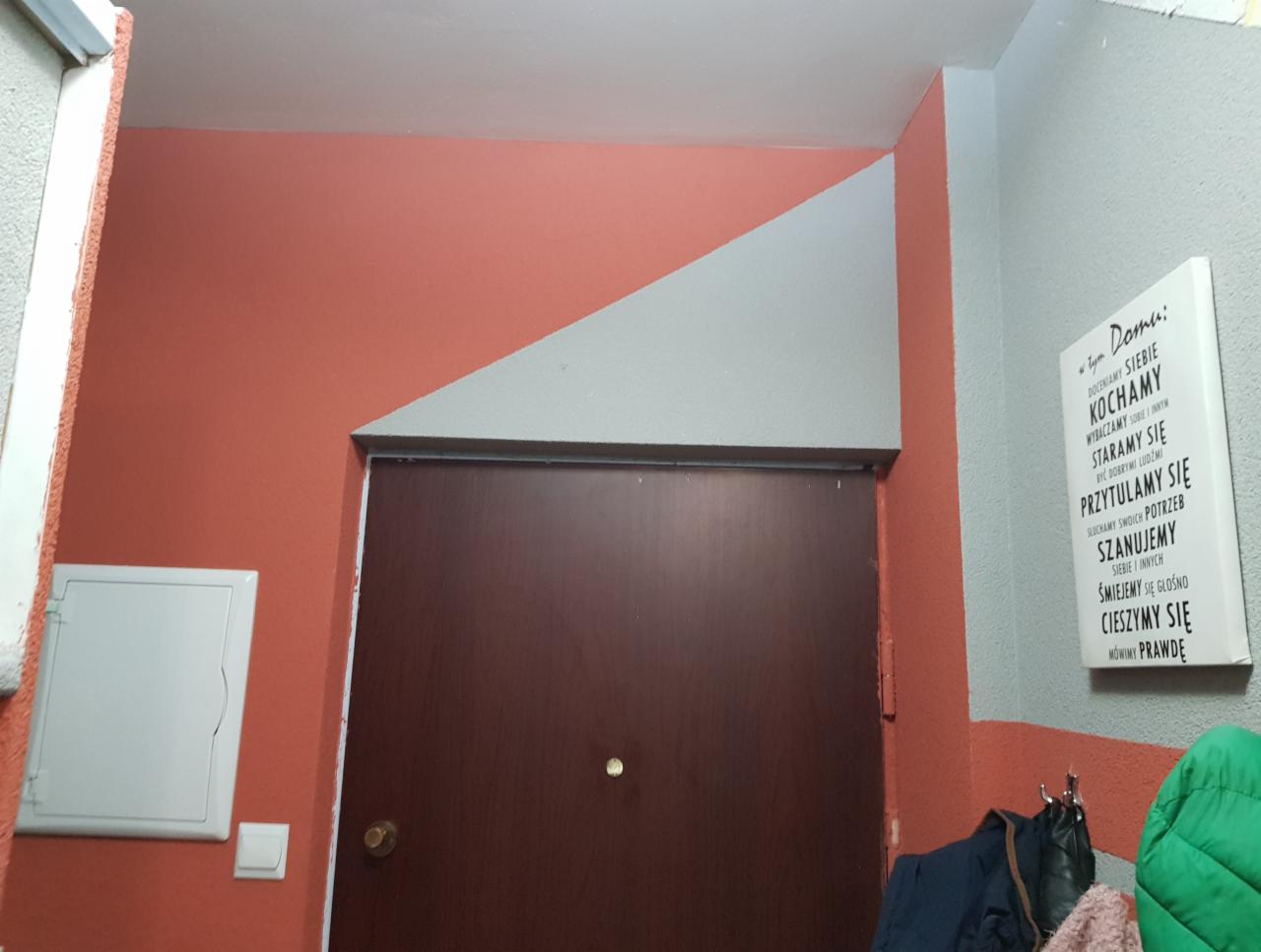 Korytarz po malowaniu: ściany w kolorze terakoty i szarości, z dekoracyjnym trójkątnym elementem nad brązowymi drzwiami. Na ścianie wisi biała tabliczka z napisem 'W tym domu...'