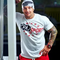 Uśmiechnięty mężczyzna w sportowym stroju, oparty o maszynę na siłowni, prezentuje tatuaże na ramionach i zegarek na ręce.