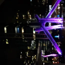 Nocny widok na oświetloną konstrukcję z neonowym fioletowym światłem i budynki w tle, ujęcie z balkonu.