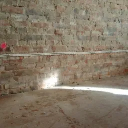 Surowa ceglana ściana z natynkowymi przewodami elektrycznymi i puszkami instalacyjnymi, widoczne ślady wiercenia i r&oacute;żowy punkt na ścianie, betonowa podłoga z plamami.