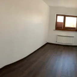 Pusty, odświeżony pokój z ciemną podłogą imitującą drewno, białymi ścianami, grzejnikiem pod oknem z brązową ramą i roletami plisowanymi, oraz białą, nowoczesną stolarką drzwiową.