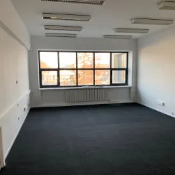 Puste pomieszczenie biurowe z dużym oknem, świeżo pomalowane ściany na biało, ciemna wykładzina dywanowa, widoczne grzejniki pod oknem i listwy przypodłogowe.