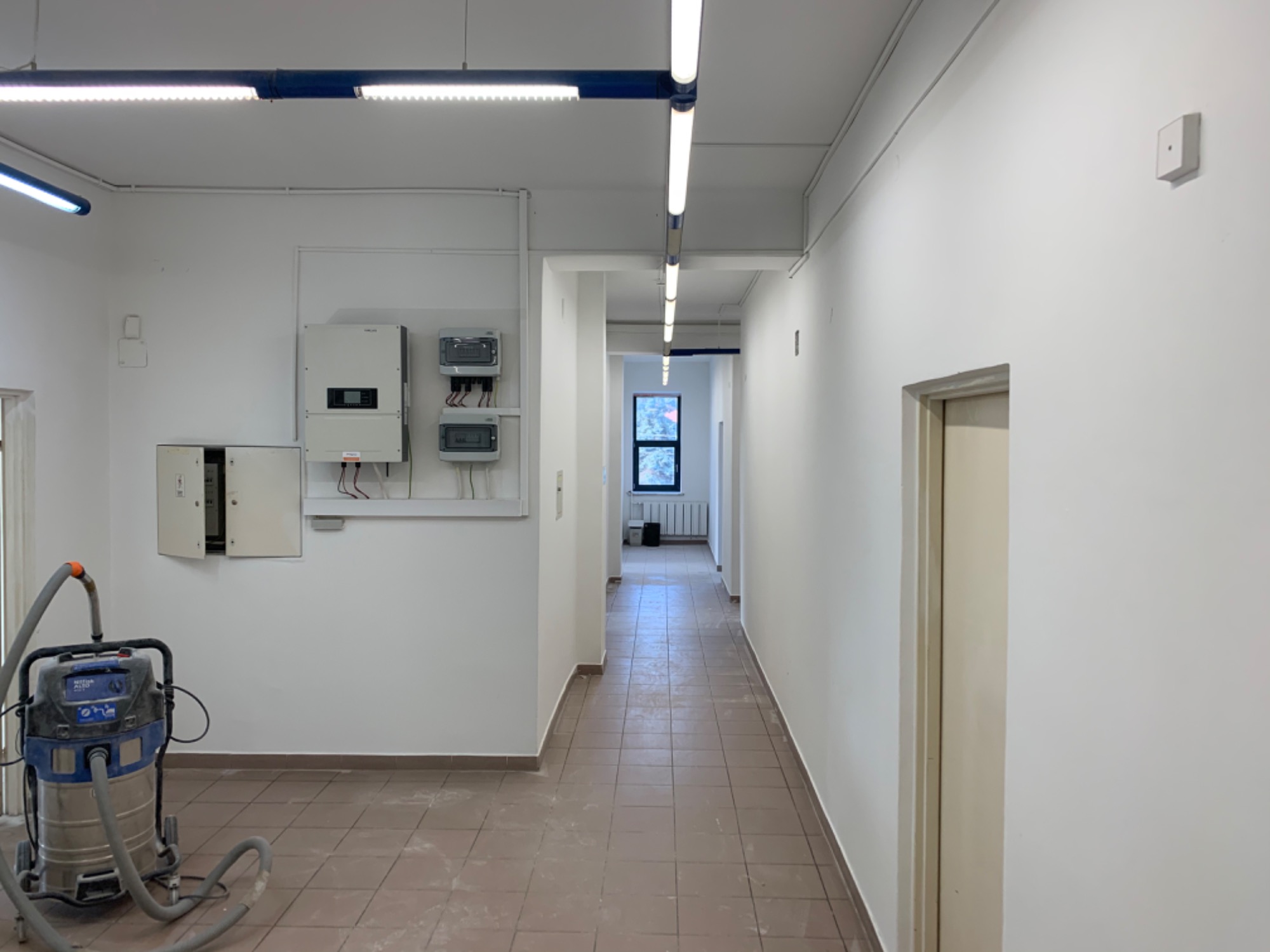 Korytarz biurowy po remoncie, widoczny odkurzacz przemysłowy, świeżo pomalowane ściany na biało, brązowa płytka podłogowa i instalacje elektryczne.