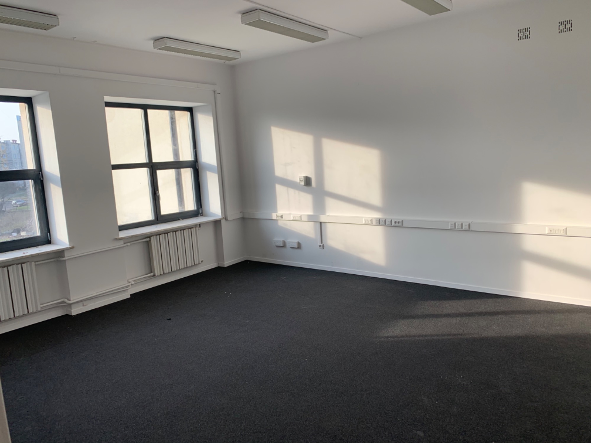 Puste, jasne biuro z dwoma oknami i ciemną wykładziną dywanową. Świeże, białe ściany z listwami przypodłogowymi i gniazdkami elektrycznymi. Widoczne grzejniki pod oknami.