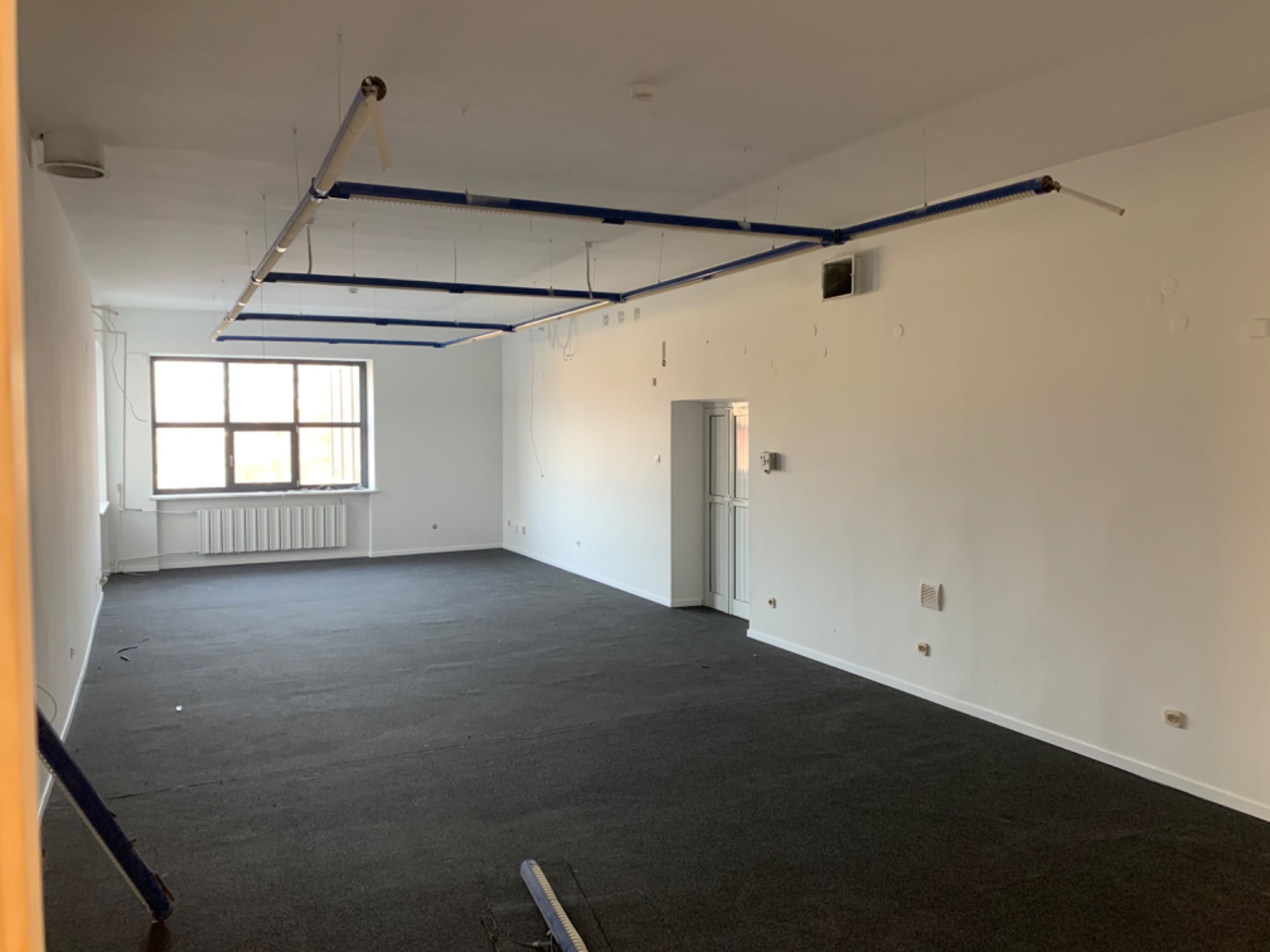 Wnętrze biura z ciemną wykładziną dywanową, białymi ścianami z widocznymi śladami po instalacjach i oświetleniem sufitowym z niebieskimi elementami konstrukcyjnymi.