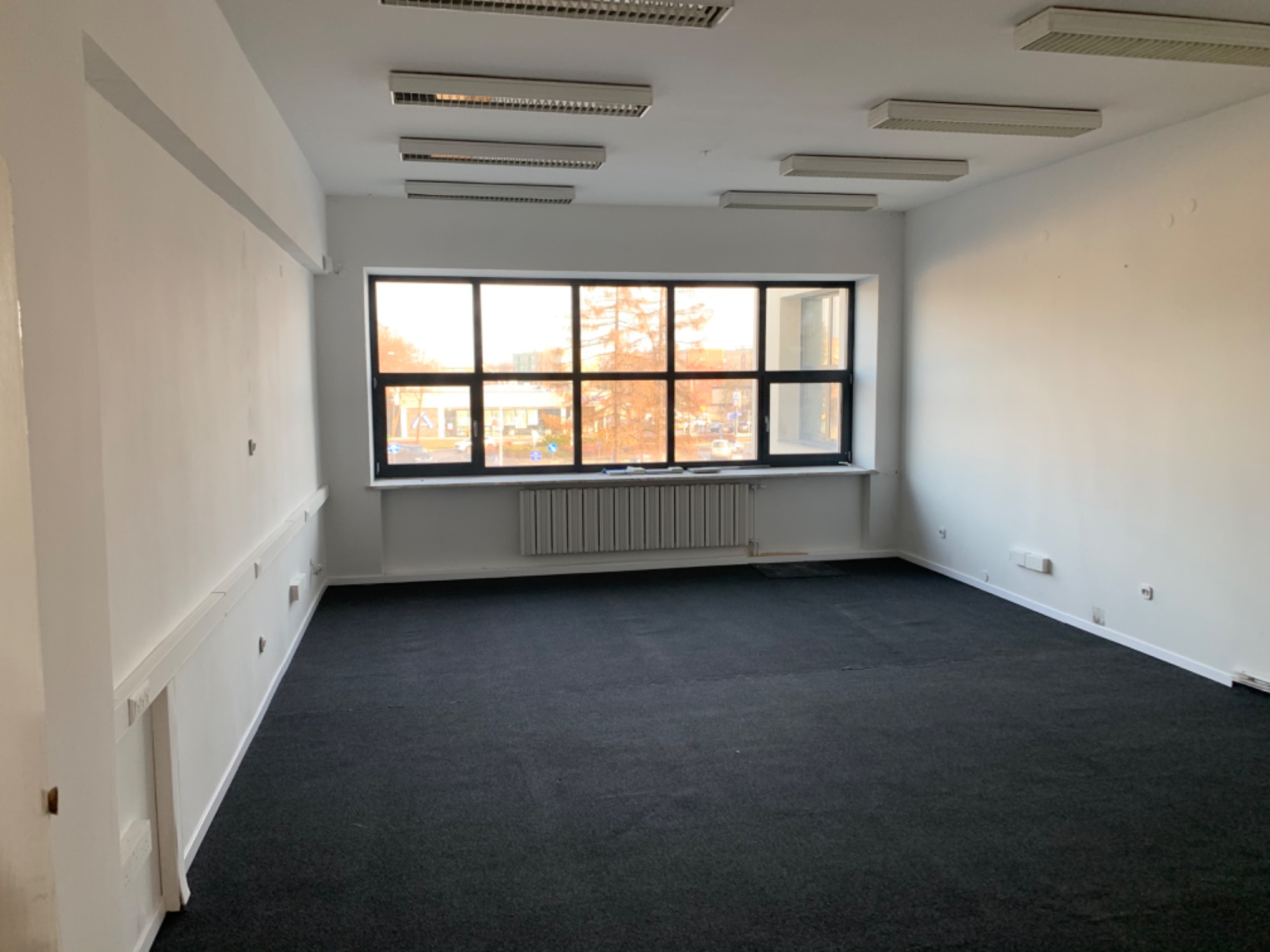Puste pomieszczenie biurowe z dużym oknem, świeżo pomalowane ściany na biało, ciemna wykładzina dywanowa, widoczne grzejniki pod oknem i listwy przypodłogowe.