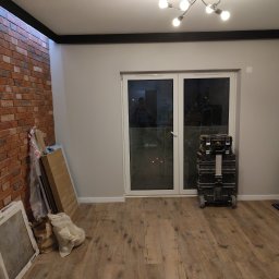 RemVik - Pokój po częściowym remoncie: ułożona podłoga drewniana, widoczna ściana z cegły, narzędzia i materiały budowlane przy ścianie, białe drzwi balkonowe.