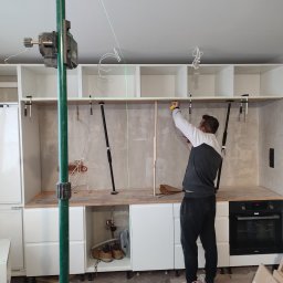 RemVik - Pracownik w Łodzi mierzy odległość między szafkami kuchennymi za pomocą miarki. Widoczny laser krzyżowy, szafki bez frontów, sprzęt AGD i narzędzia.