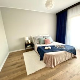 RemVik - Jasna sypialnia z drewnianą podłogą, szarą ścianą i łóżkiem z niebieską narzutą oraz dekoracyjnymi poduszkami. Widoczny stolik nocny z lampką i otwartą książką na łóżku. Z okna wpada naturalne...