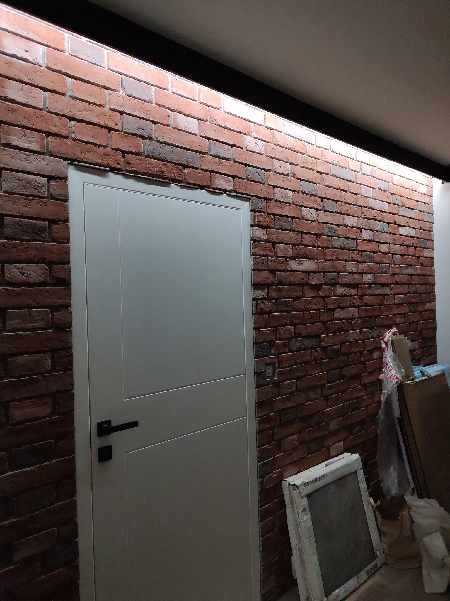 Białe drzwi z czarną klamką na tle ściany z cegły klinkierowej z podświetleniem LED u góry, w tle materiały budowlane.