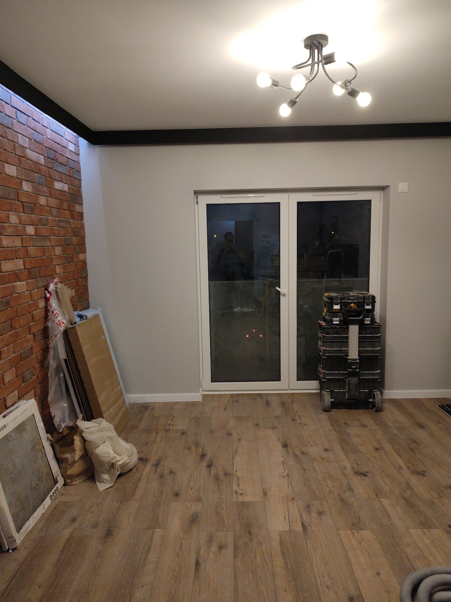 Pokój po częściowym remoncie: ułożona podłoga drewniana, widoczna ściana z cegły, narzędzia i materiały budowlane przy ścianie, białe drzwi balkonowe.