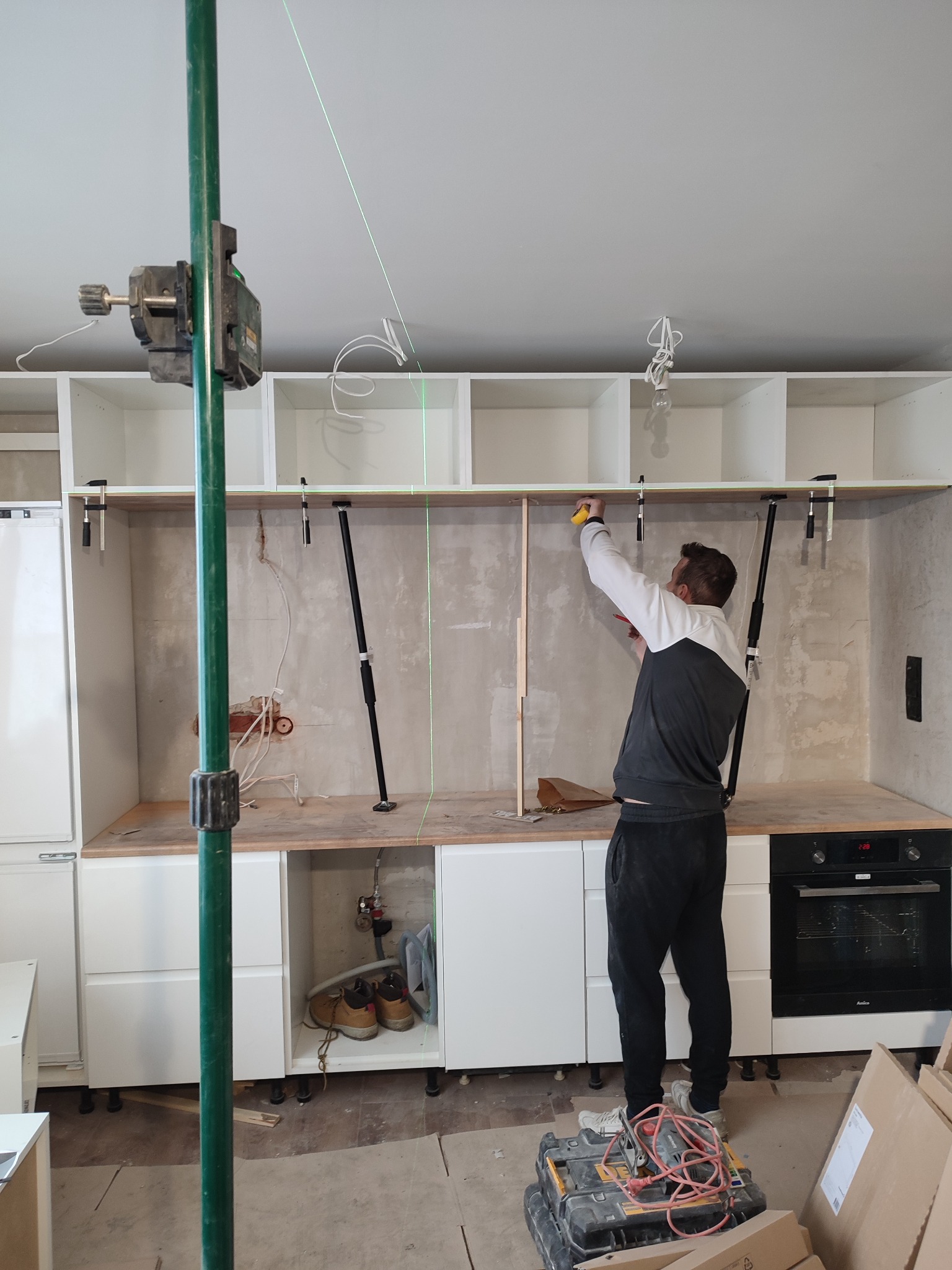 Pracownik w Łodzi mierzy odległość między szafkami kuchennymi za pomocą miarki. Widoczny laser krzyżowy, szafki bez frontów, sprzęt AGD i narzędzia.