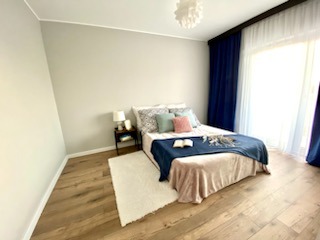 Jasna sypialnia z drewnianą podłogą, szarą ścianą i łóżkiem z niebieską narzutą oraz dekoracyjnymi poduszkami. Widoczny stolik nocny z lampką i otwartą książką na łóżku. Z okna wpada naturalne...